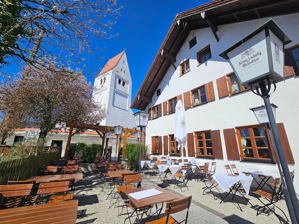 An outdoor seating area with tables and chairs, a white building with wooden shutters, and a bell tower.