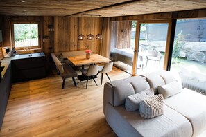 A modern interior with a wooden floor, a dining area with a table and chairs, a sofa, and a large window offering a view of the outdoors. - Slow Fine Chalets (Neustift Im Stubaital)
