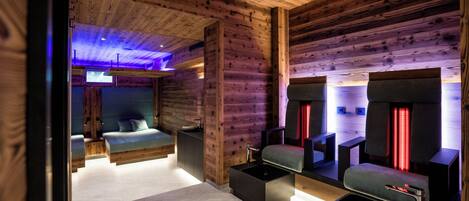 Interior de una cabaña rústica moderna con una cama, un baño con espejo y luces rojas, y un pequeño rincón de descanso.
