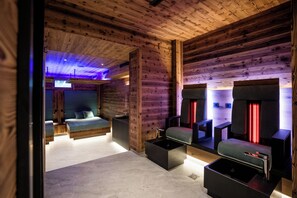 Interior de una cabaña rústica moderna con una cama, un baño con espejo y luces rojas, y un pequeño rincón de descanso.