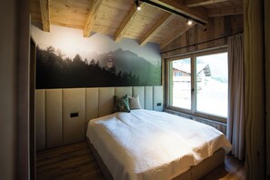 A bedroom with a large bed, a wooden ceiling, a mountain mural, and a window with curtains.
