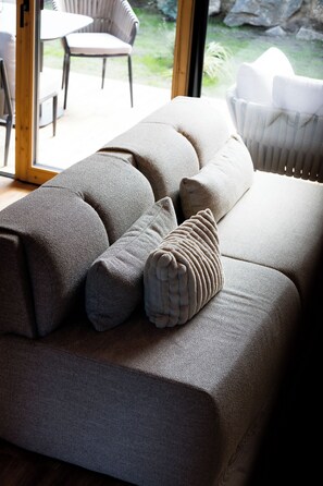 A modern living room with a large grey sofa, a patterned throw pillow, and a view of the outdoors through a glass door.