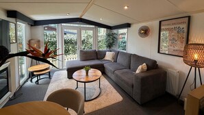 A modern living room with a grey sofa, round coffee table, and a large window providing a view of greenery outside. - Chalet with Jacuzzi and Private Garden - Shared Pool in Summer Season (Hoenderloo)