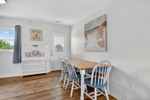 A dining area with a wooden table and white chairs, a door with a sign, and a painting on the wall. - UNIT A: Walk to Beach/Intercoastal/Fishing Pier (Holly Ridge)