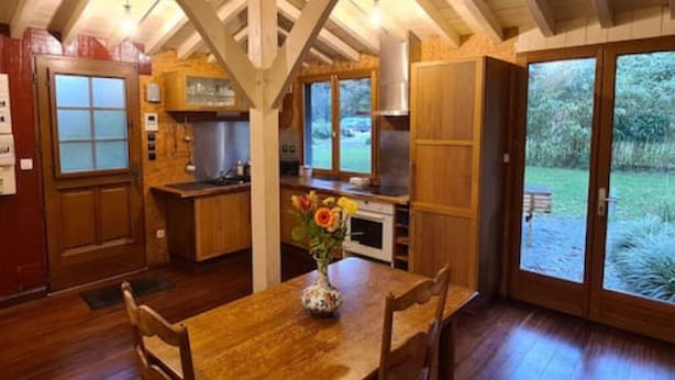 Un interior de madera con una mesa de comedor, sillas, un jarrón con flores y una zona de cocina con armarios y una ventana.