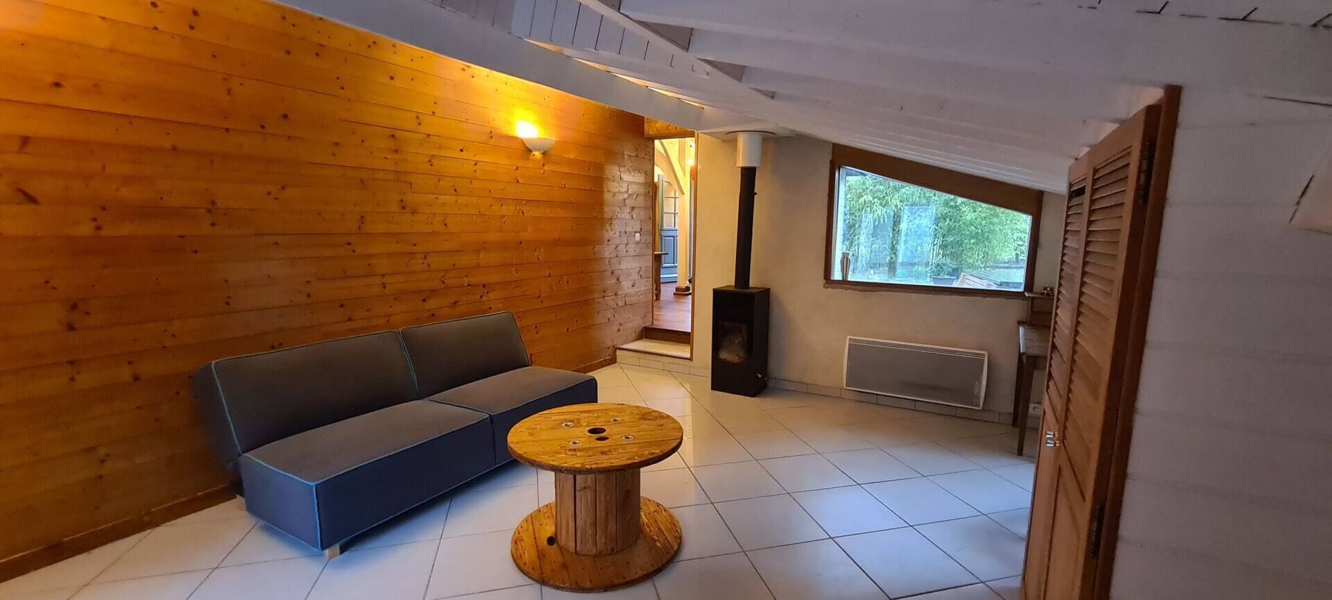Ein Zimmer mit einer Holzwand, einem blauen Sofa, einem runden Holztisch, einem schwarzen Lautsprecher und einem Fenster mit Blick auf Grünflächen.