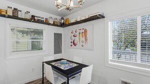Ein Essbereich mit Tisch, Stühlen, einer Chandelieber, einem Fenster mit Jalousien und einem an der Wand befestigten Regal mit Gläsern.