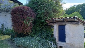 Een rustiek stenen gebouw met een klein aanbouwtje, omgeven door een tuin met diverse planten en een grindpad.