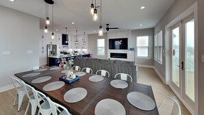 Una cocina moderna con armarios blancos, una zona de comedor con una gran mesa de madera y sillas blancas, y un televisor montado en una pared de piedra.