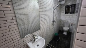 Une salle de bain avec des murs recouverts de carreaux blancs, un lavabo blanc, une douche et des toilettes.