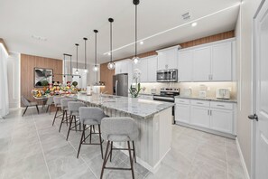 Eine moderne Küche mit weißen Schränken, einer großen Kochinsel mit Barhocker, Edelstahlgeräten, Pendelleuchten und einer Wand aus Holzpaneelen.
