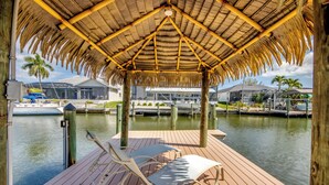 A wooden deck with a thatched roof, overlooking calm water with boats and buildings in the background. - Tropical Tiki, Heated Pool, Waterfront Gulf Access, Dock your boat, WIFI (Cape Coral)