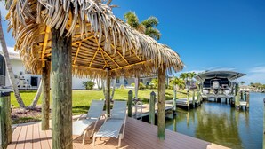 A wooden deck with lounge chairs, a thatched roof structure, a water body, and a dock with a boat. - Tropical Tiki, Heated Pool, Waterfront Gulf Access, Dock your boat, WIFI (Cape Coral)