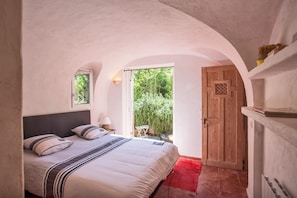 Ein Schlafzimmer mit einem Bett, einem Nachttisch, einer Lampe, einer Holztür und Blick ins Grüne.