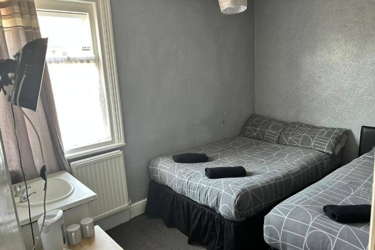 Ein kleines Zimmer mit einem Bett, einem Fenster, einem Waschbecken und einer Bettdecke mit geometrischem Muster.