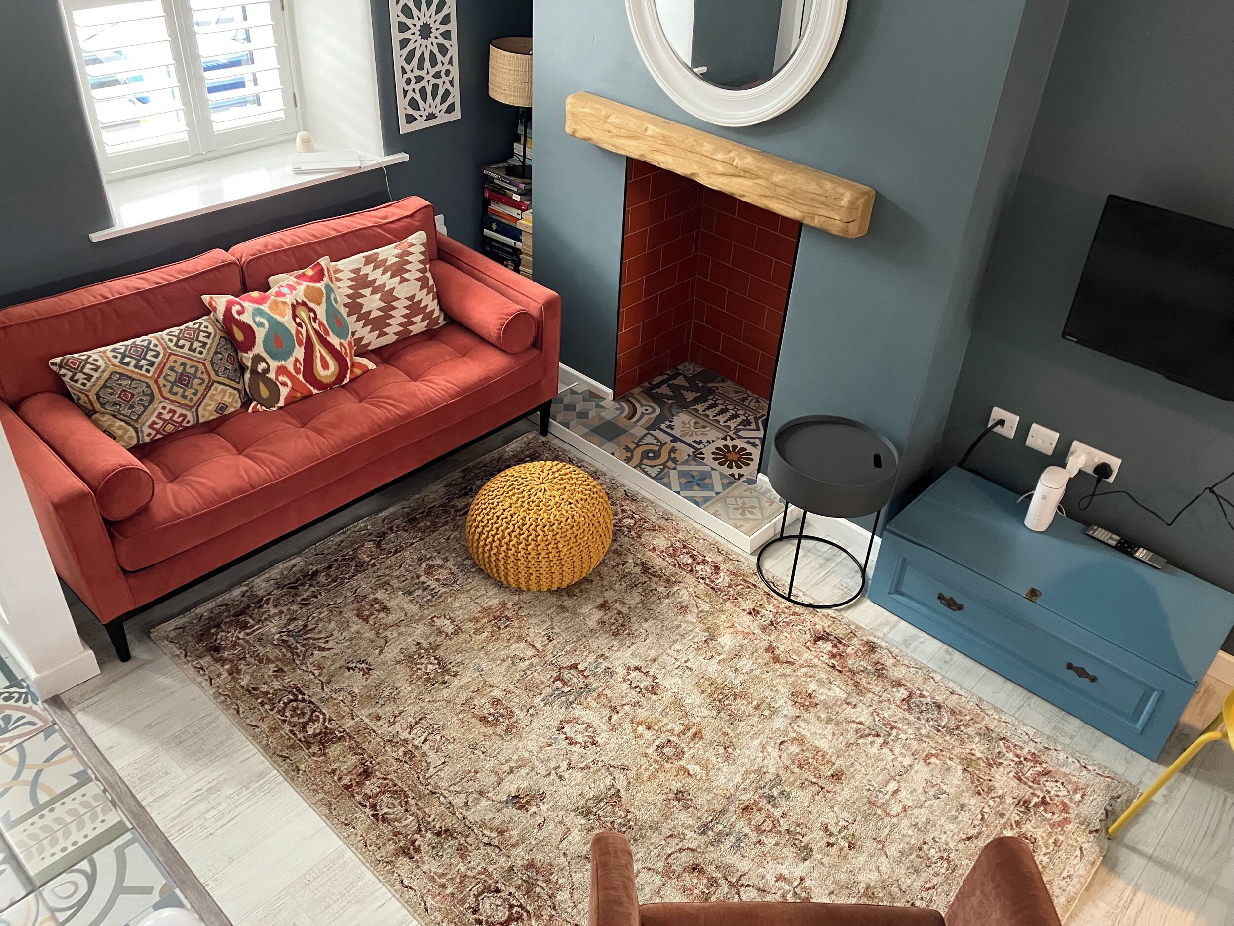 A living room with a red sofa, a patterned rug, a round yellow ottoman, a blue cabinet, and a fireplace.