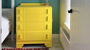 A room with a yellow chest of drawers, a white bed, a red-framed mirror, and a lamp on a wooden stand. - East Meets West (Criccieth)