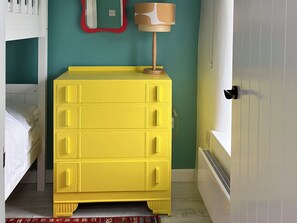 A room with a yellow chest of drawers, a white bed, a red-framed mirror, and a lamp on a wooden stand. - East Meets West (Criccieth)