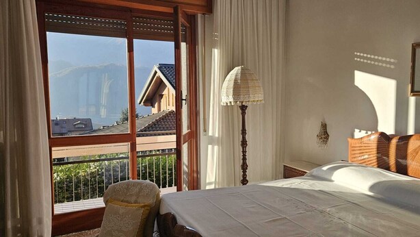 Ein Schlafzimmer mit Bett, Nachttisch, Lampe, Stuhl und Blick auf Gebäude durch ein Fenster.