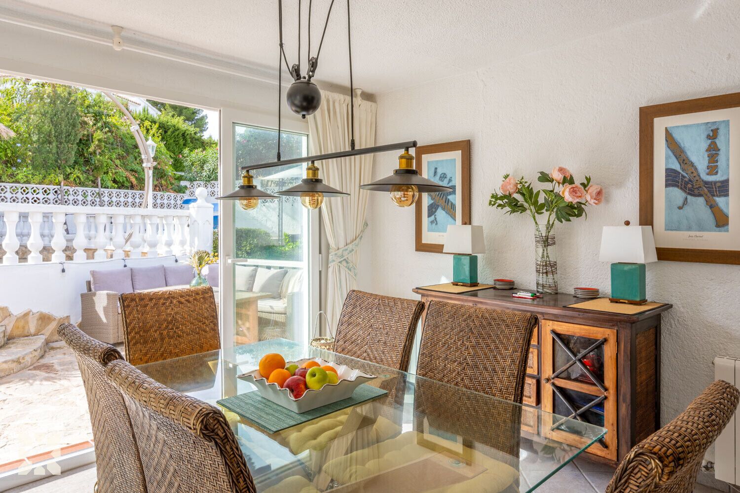 A dining area with a glass table, wicker chairs, a fruit bowl, and a wooden cabinet with a lamp.