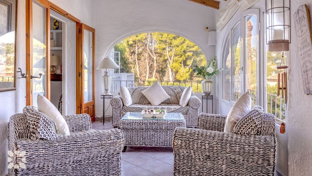 A cozy room with wicker furniture, a round table, and a view of trees through a large arched window. - Villa CATALINA by Abahana Villas (Teulada)
