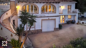 A two-story house with a white exterior, a stone base, a curved staircase, and a covered patio area. - Villa CATALINA by Abahana Villas (Teulada)