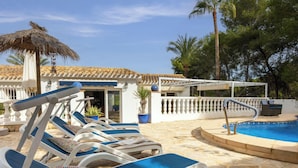 A pool area with lounge chairs, a thatched umbrella, and a building with a tiled roof. - Villa CATALINA by Abahana Villas (Teulada)