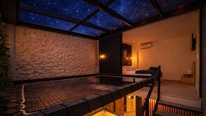 Una habitación con pared de piedra, una cama, una mesita con una botella y un vaso, y vistas del cielo estrellado a través de un techo de cristal.