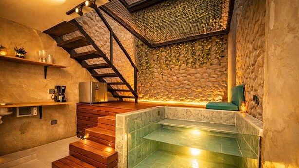 Un interior moderno con pared de piedra, escalera de madera, bañera de hidromasaje con azulejos verdes y una cafetera.