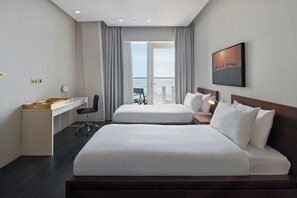 Ein Hotelzimmer mit zwei Betten, einem Schreibtisch, einem Sessel, einem Balkon mit Aussicht und einem Bild an der Wand.