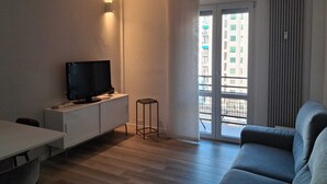 Ein modernes Wohnzimmer mit einem Flachbildfernseher auf einem weißen Schrank, einem grauen Sofa, einem kleinen Beistelltisch und einer Schiebetür zu einem Balkon.