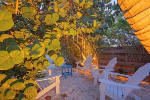 A garden area with white chairs and a blue stool, surrounded by large green leaves and a wooden fence. - Sweet Retreat Cottage: Ground level cottage across the street from the beach (Anna Maria)
