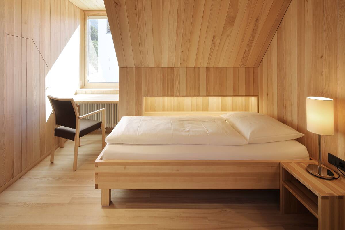 A wooden-paneled room with a bed, a chair, a desk, and a lamp.