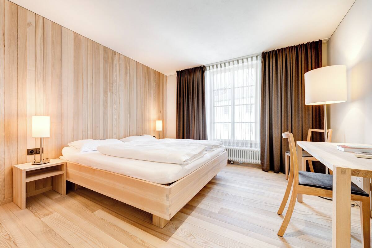 A hotel room with a wooden bed, a bedside table with a lamp, a dining table with chairs, and a large window with curtains.