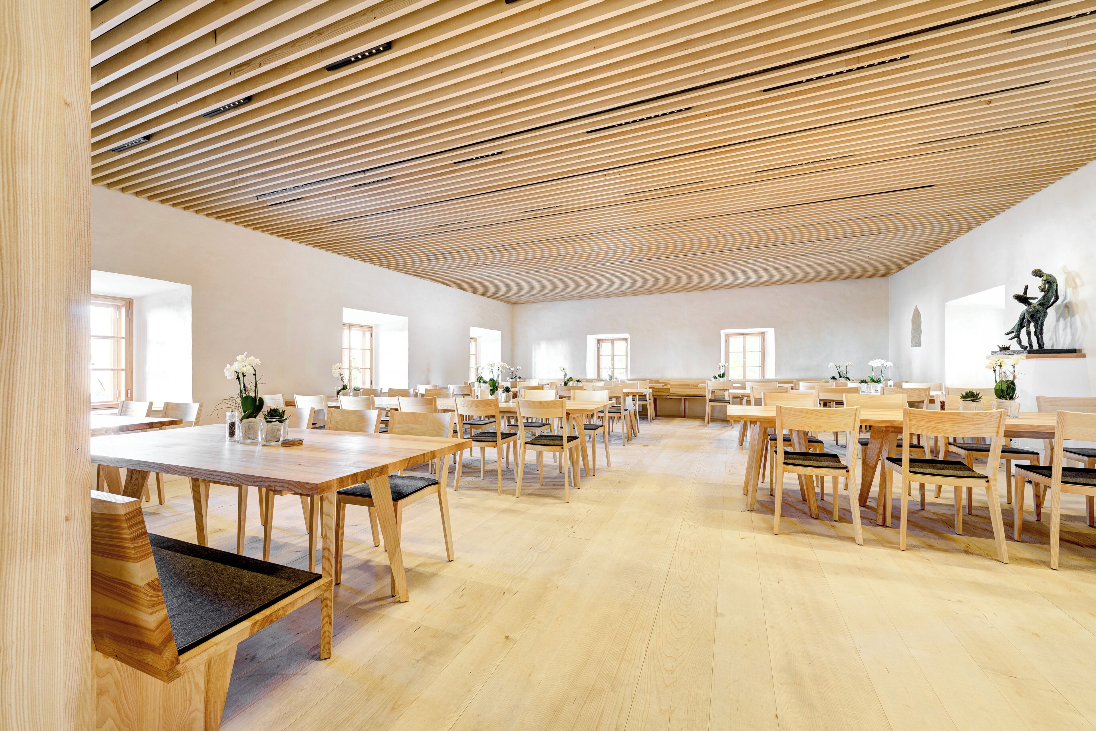 A spacious room with wooden tables and chairs, large windows, and a slatted ceiling.