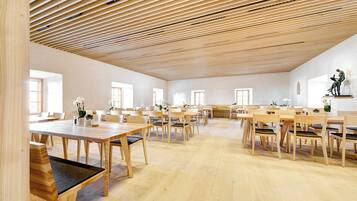 A spacious room with wooden tables and chairs, large windows, and a slatted ceiling.