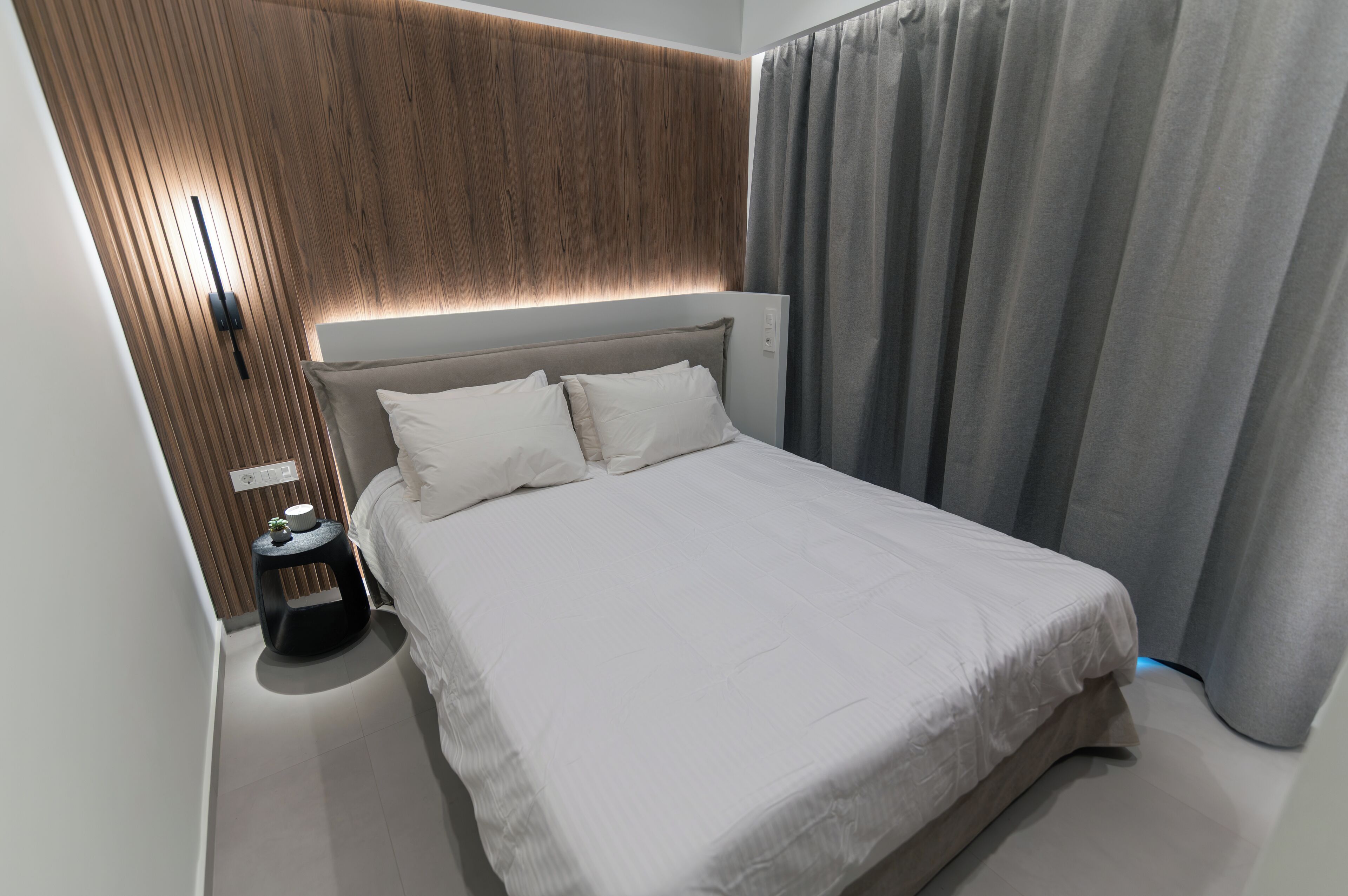 A modern hotel room with a large bed, a bedside table with a lamp, a wooden panel wall, and a curtain.