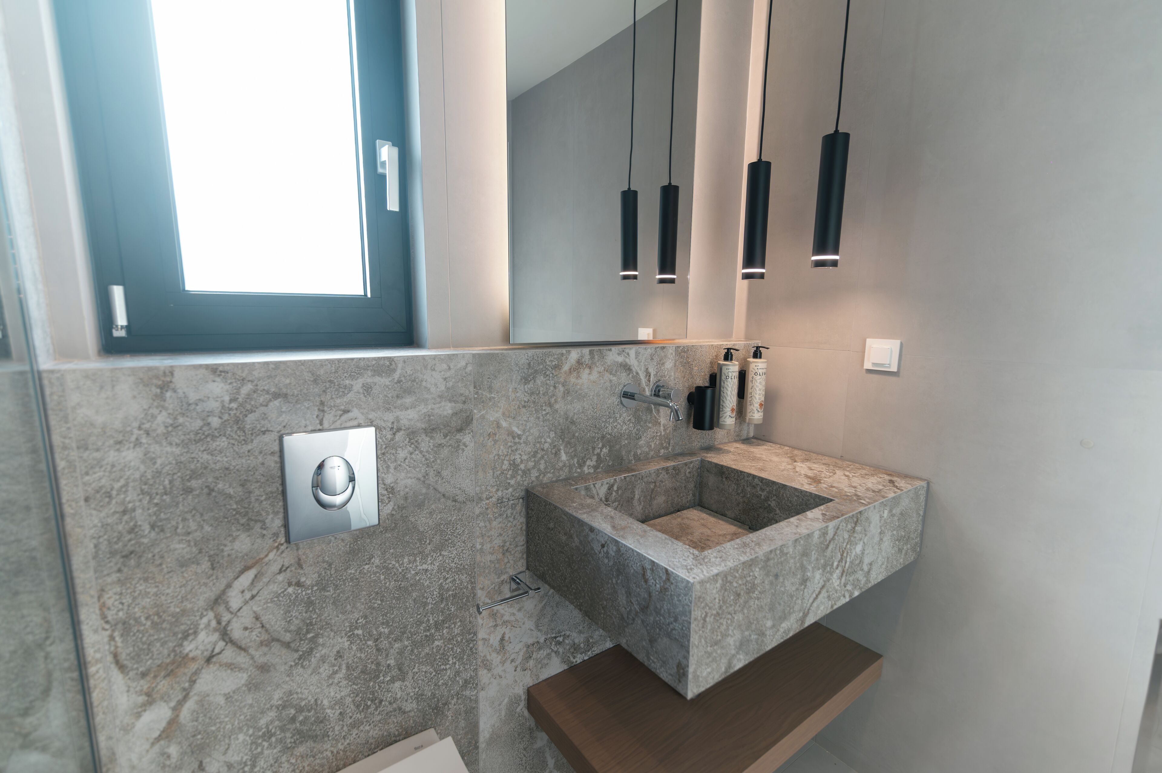 A modern bathroom with a marble sink, a mirror, pendant lights, and a window.
