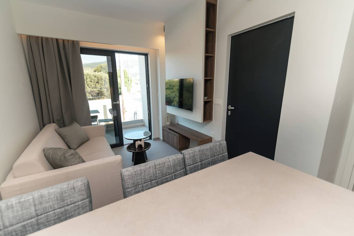 Een moderne woonkamer met een bank, een eettafel, een aan de muur bevestigde televisie en een schuifdeur naar een buitenruimte.