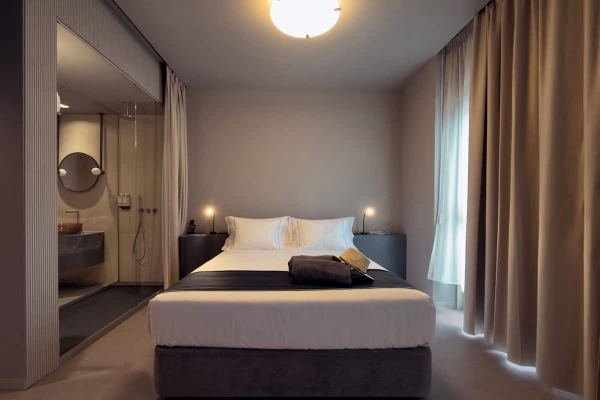Une chambre d’hôtel moderne avec un grand lit, des tables de chevet, un miroir et une salle de bain équipée d’un lavabo et d’une douche.