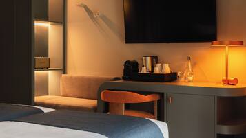 A hotel room with a bed, a desk with a lamp, a television, and a seating area with a chair.