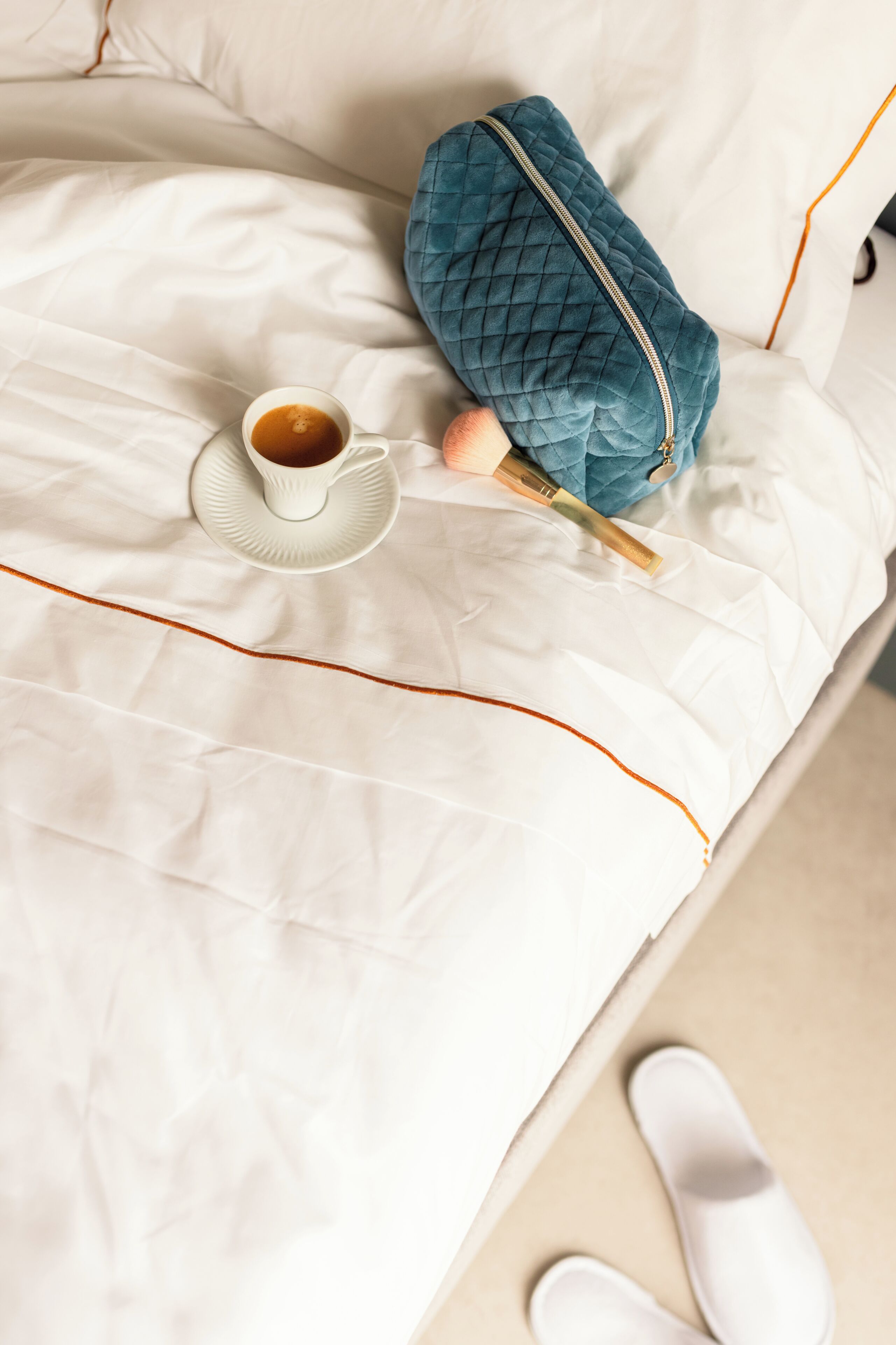 A neatly made bed with a white sheet, a cup of coffee, a blue quilted bag, and a pair of slippers.