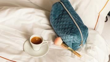 A neatly made bed with a white sheet, a cup of coffee, a blue quilted bag, and a pair of slippers.