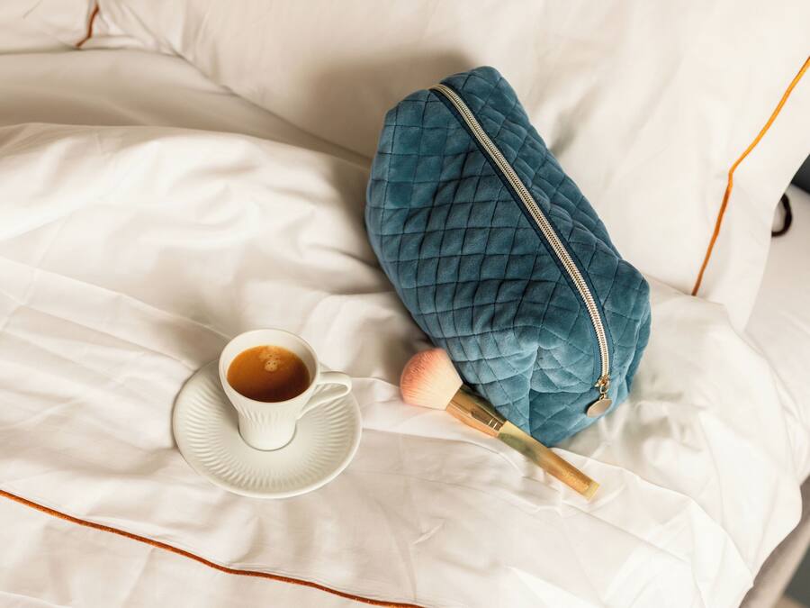 A neatly made bed with a white sheet, a cup of coffee, a blue quilted bag, and a pair of slippers.