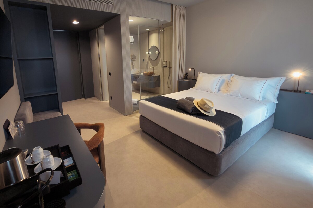 A modern hotel room with a large bed, a desk with a tray of breakfast items, a shower area, and a mirror.