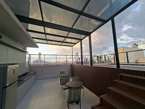A modern balcony with a glass ceiling, a dining area with chairs, and a view of the city skyline. - Estúdio e lazer completo piscina rooftop (São Paulo)