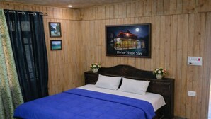 A bedroom with a wooden paneled wall, a bed with a blue bedspread, two framed pictures, and a sign above the bed. - Divine Home stay  (Rajgarhi)