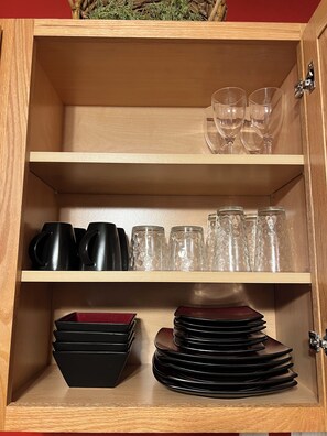 A wooden cabinet with shelves containing black mugs, clear glasses, and stacked black plates.