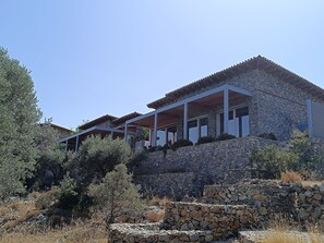 Ein Gebäude aus Stein mit einer flachen Dachterrasse und zahlreichen Fenstern, umgeben von trockener Vegetation und Bäumen.