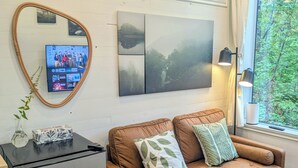Een gezellige kamer met een bruine bank, een kleine koelkast, een flatscreen-tv aan een witte muur en een groot raam met uitzicht op bomen.